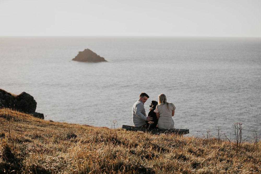 A couple sits with their dog on a clifftop bench overlooking a calm ocean with a small rocky island.
