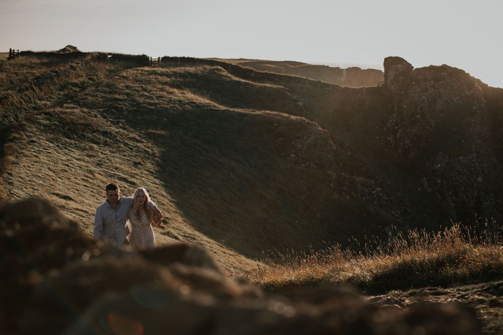 A couple holds hands while walking along a dramatic hillside path bathed in warm golden sunlight.