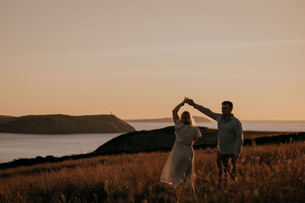 A couple dances together at sunset in a golden coastal meadow with stunning ocean and headland views.