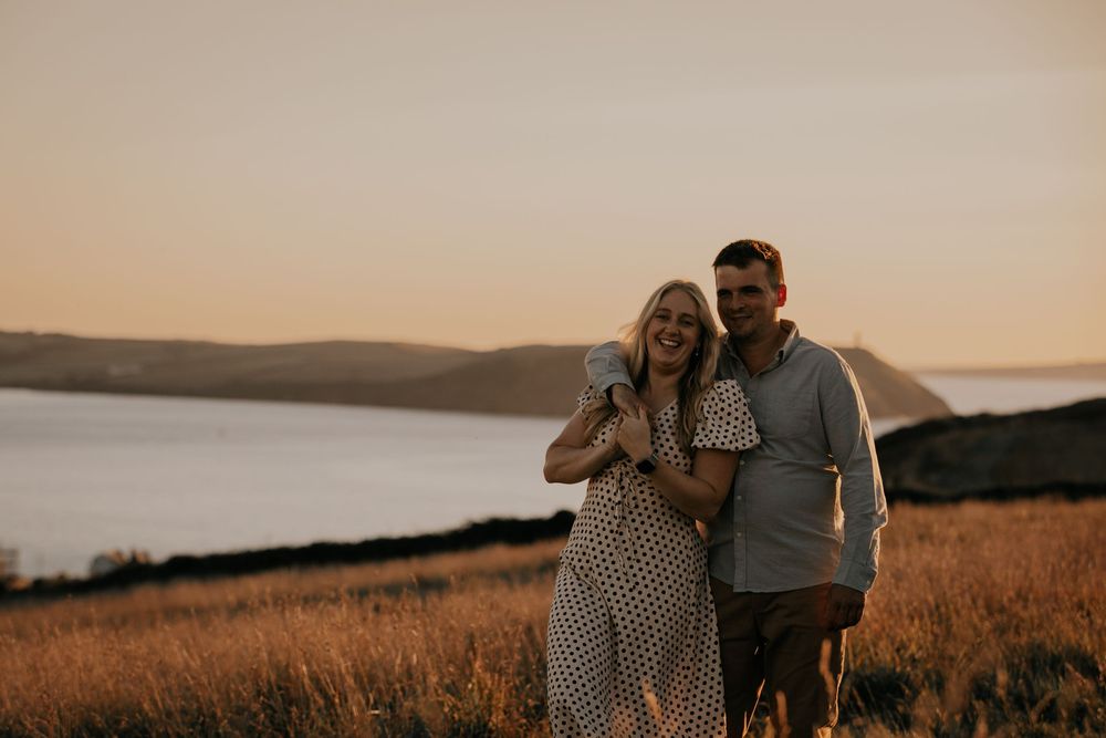 A smiling couple embraces in a golden field at sunset with a scenic coastal estuary behind them.
