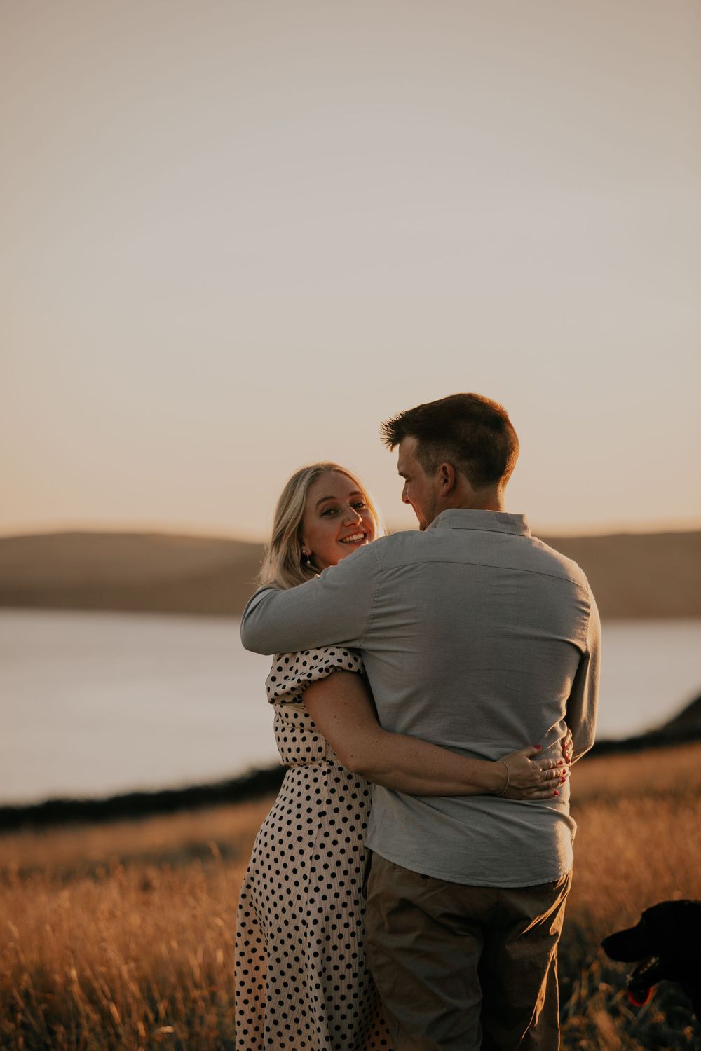 A woman in a polka dot dress smiles while embracing her partner in a golden field at sunset.