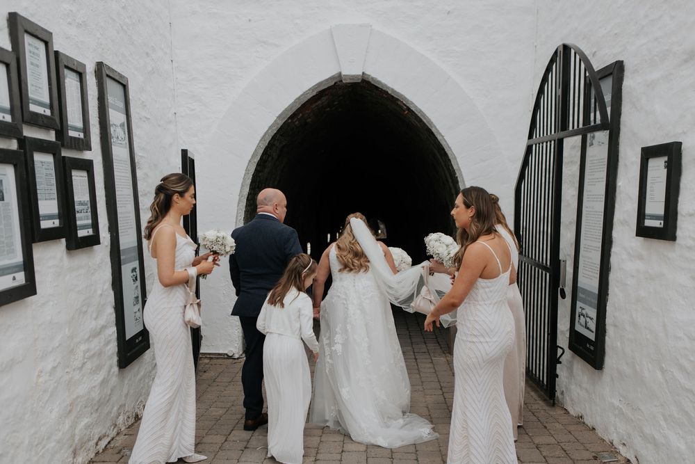 A bride with bridesmaids and flower girls entering a white stone chapel through an arched doorway.