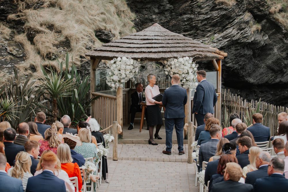 An outdoor wedding ceremony beneath a floral gazebo with seated guests and rocky cliffs behind.
