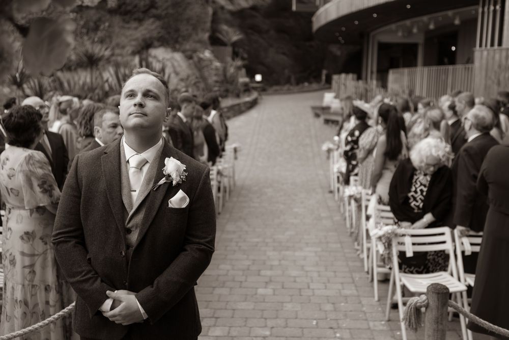 Black and white photo of a groom looking up emotionally at an outdoor wedding ceremony aisle.