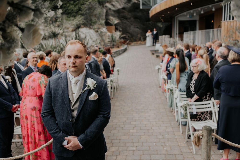 A groom in a navy suit stands at the end of the aisle awaiting his bride outdoors.