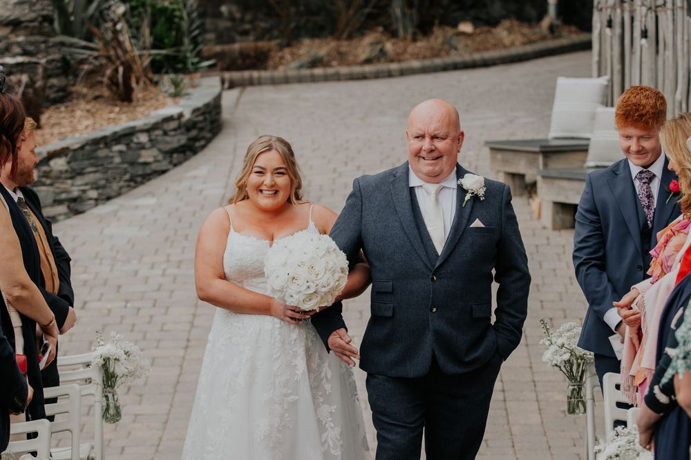 A smiling bride in a white gown walks down the aisle with her father at an outdoor wedding.