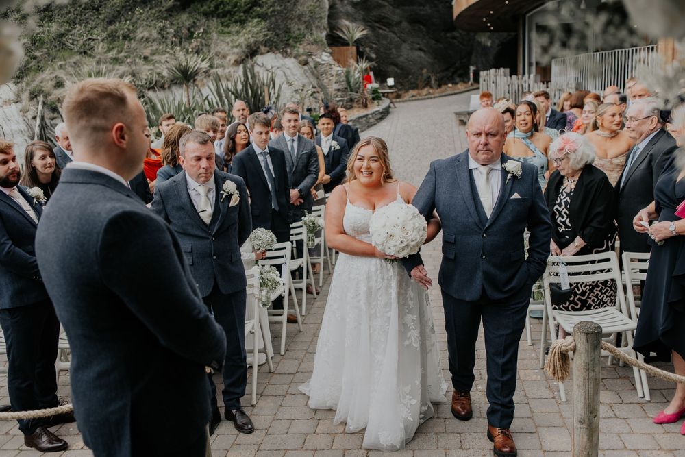 A bride walks down the aisle with her father toward the groom at an outdoor wedding ceremony.