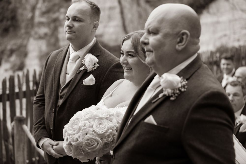Black and white photo of bride holding white rose bouquet walking down the aisle with groom and father at outdoor wedding.