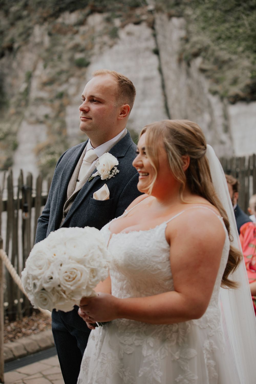 Smiling bride in lace gown holding white rose bouquet stands beside groom in navy suit at coastal wedding ceremony.