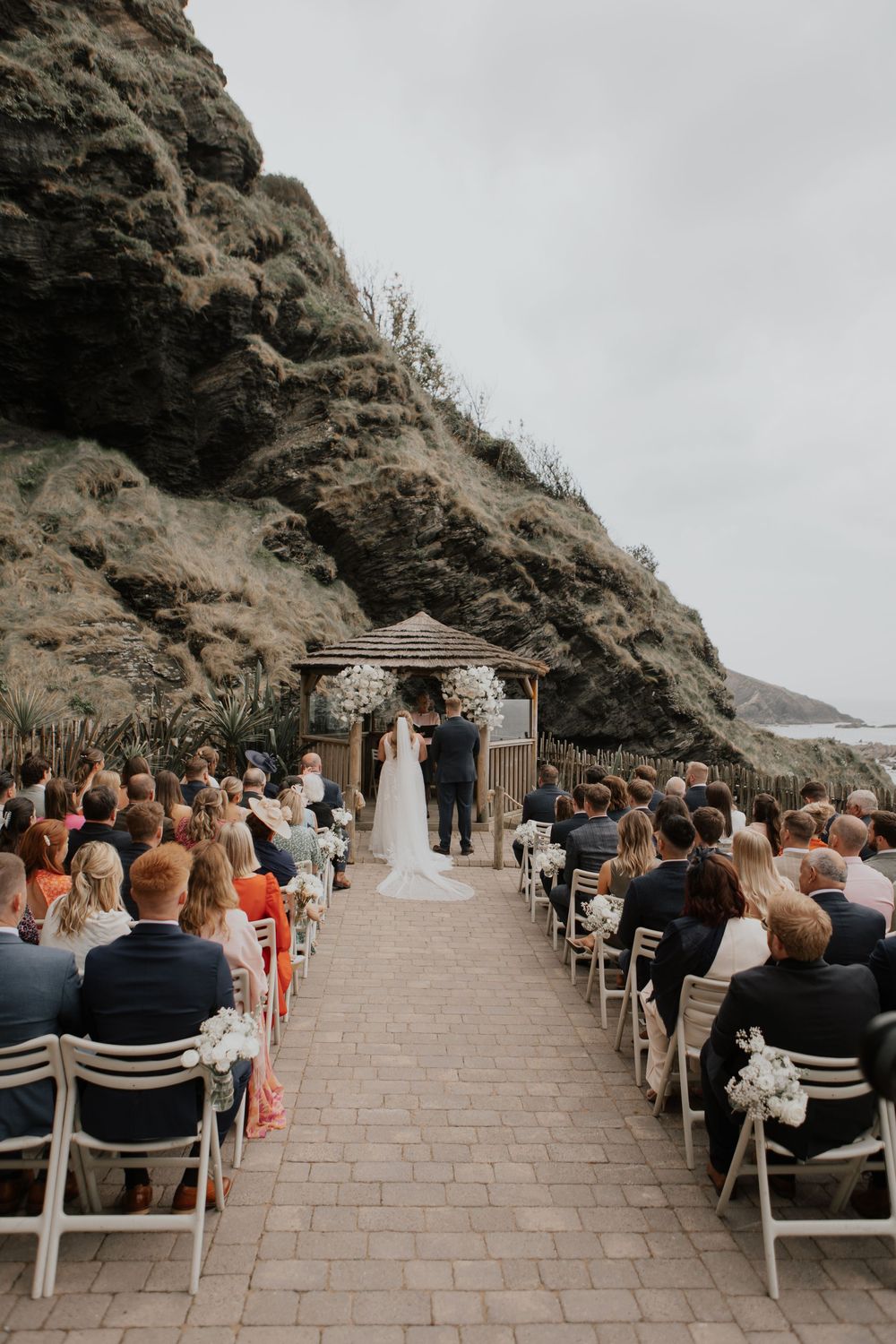 Guests seated in white chairs watch bride and groom at elegant outdoor coastal wedding ceremony beside dramatic cliffs.