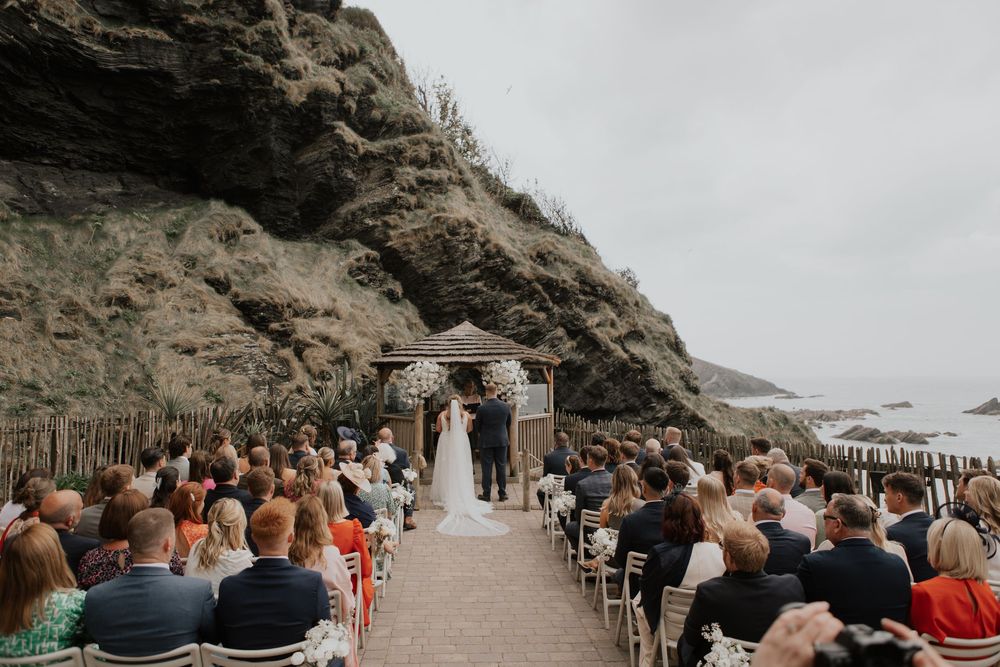Wide shot of outdoor seaside wedding ceremony with guests seated in rows facing couple at floral decorated gazebo altar.