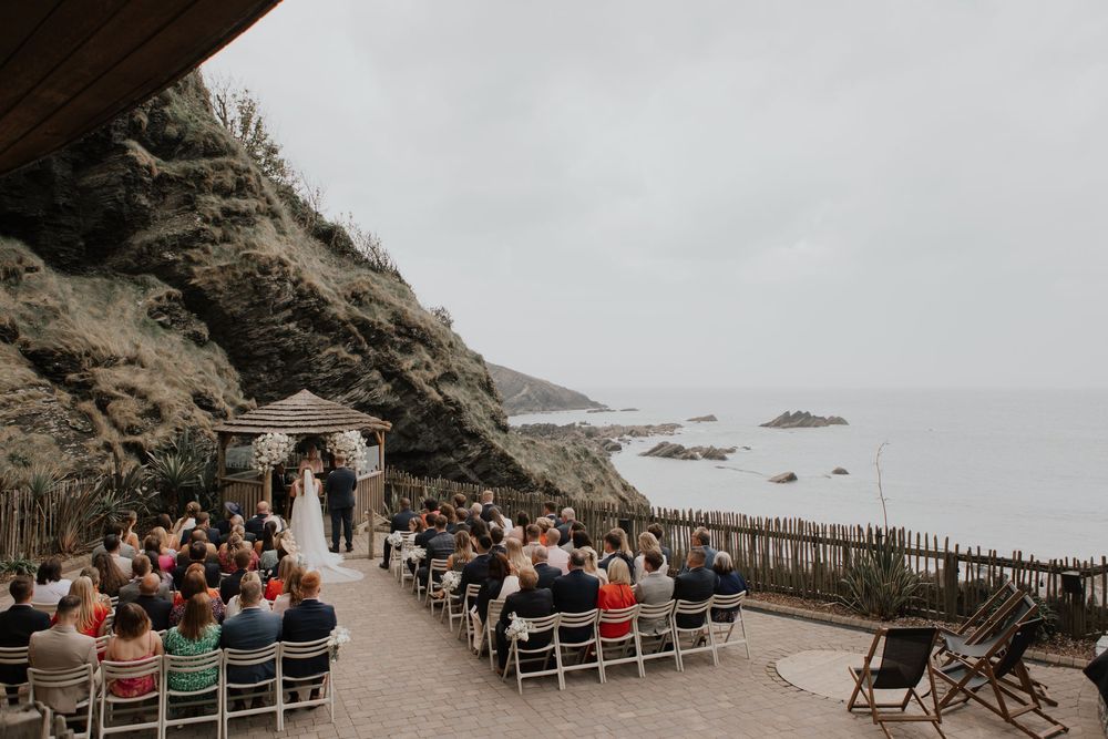 Aerial view of romantic outdoor cliff wedding ceremony with ocean backdrop and guests seated in white chairs.