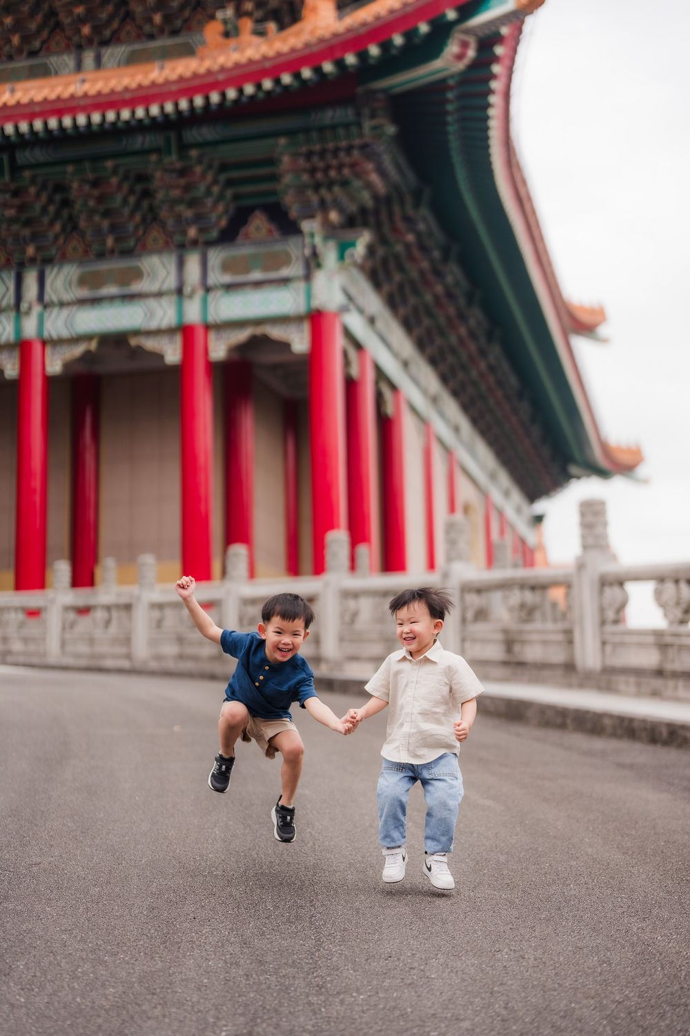 Two young Asian boys jump and pose playfully in front of traditional Chinese architecture with red pillars and ornate details.