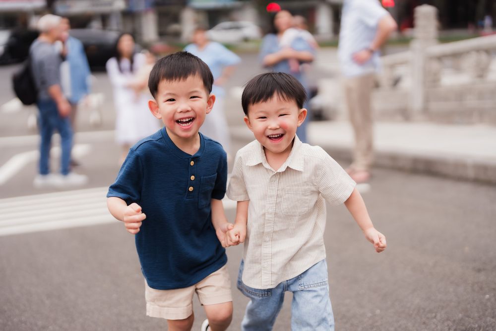 Two young boys running and playing together outdoors with joyful expressions on their faces as their families stand at a distance.