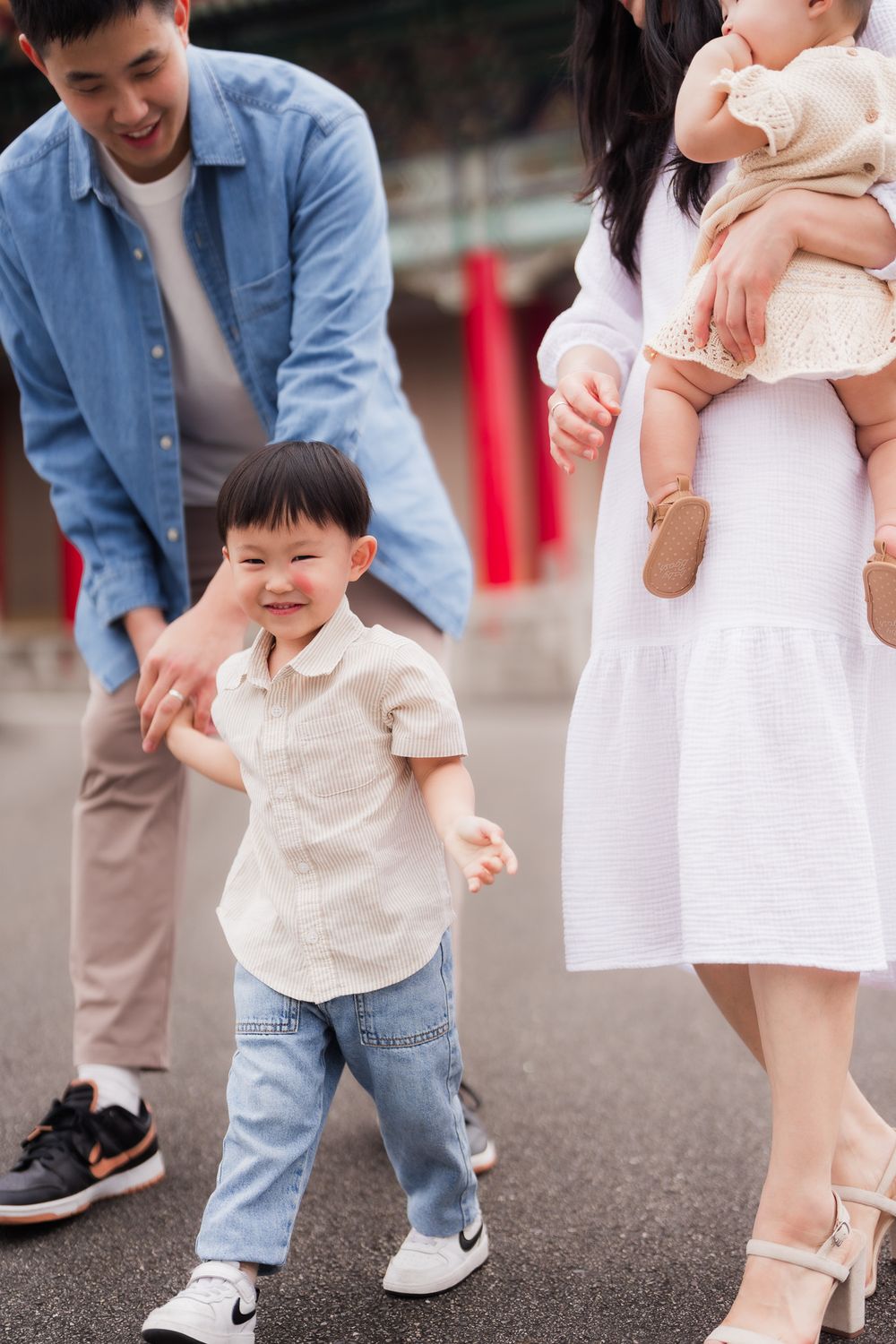 Artistic shot of a family enjoying time together outdoors with young child in beige shirt and jeans taking steps while parent holds baby.