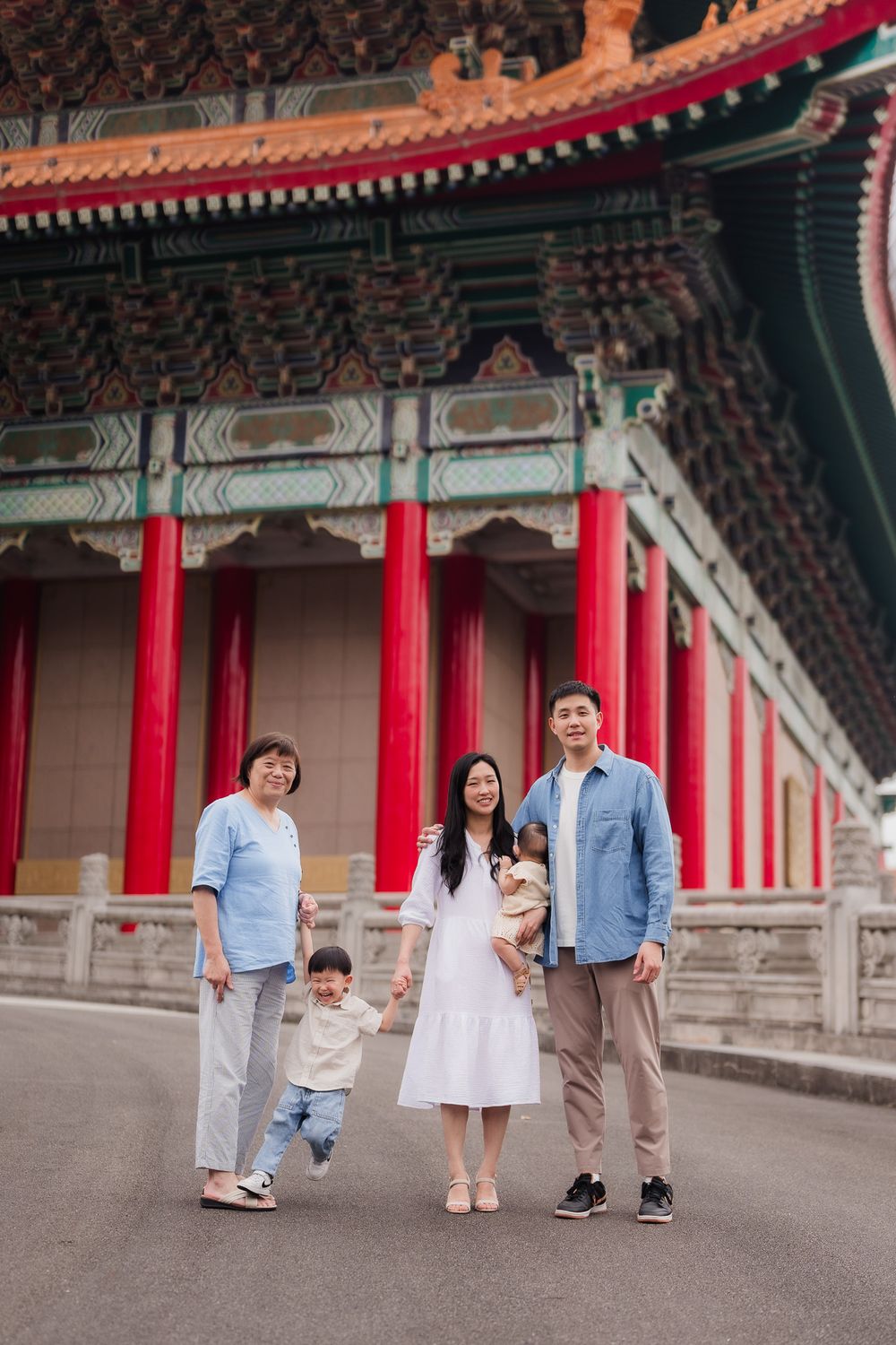 A family and grandparent pose playfully together in front of traditional Chinese architecture with red pillars and ornate rooflines.