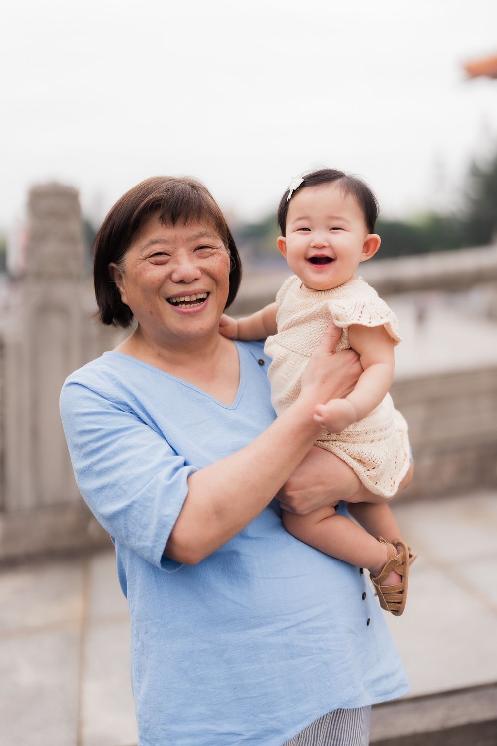 A joyful moment captured of a grandmother holding her young granddaughter outdoors with Asian style cement details in the background.