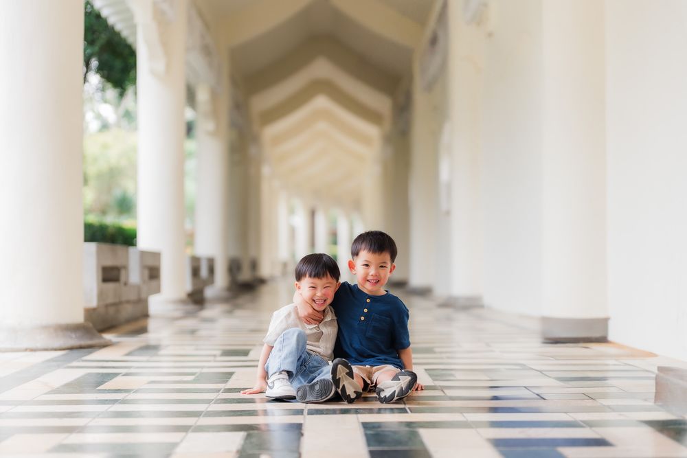 Two young boys laugh and play together on blue geometric tiles while sitting in a bright white colonnade walkway.