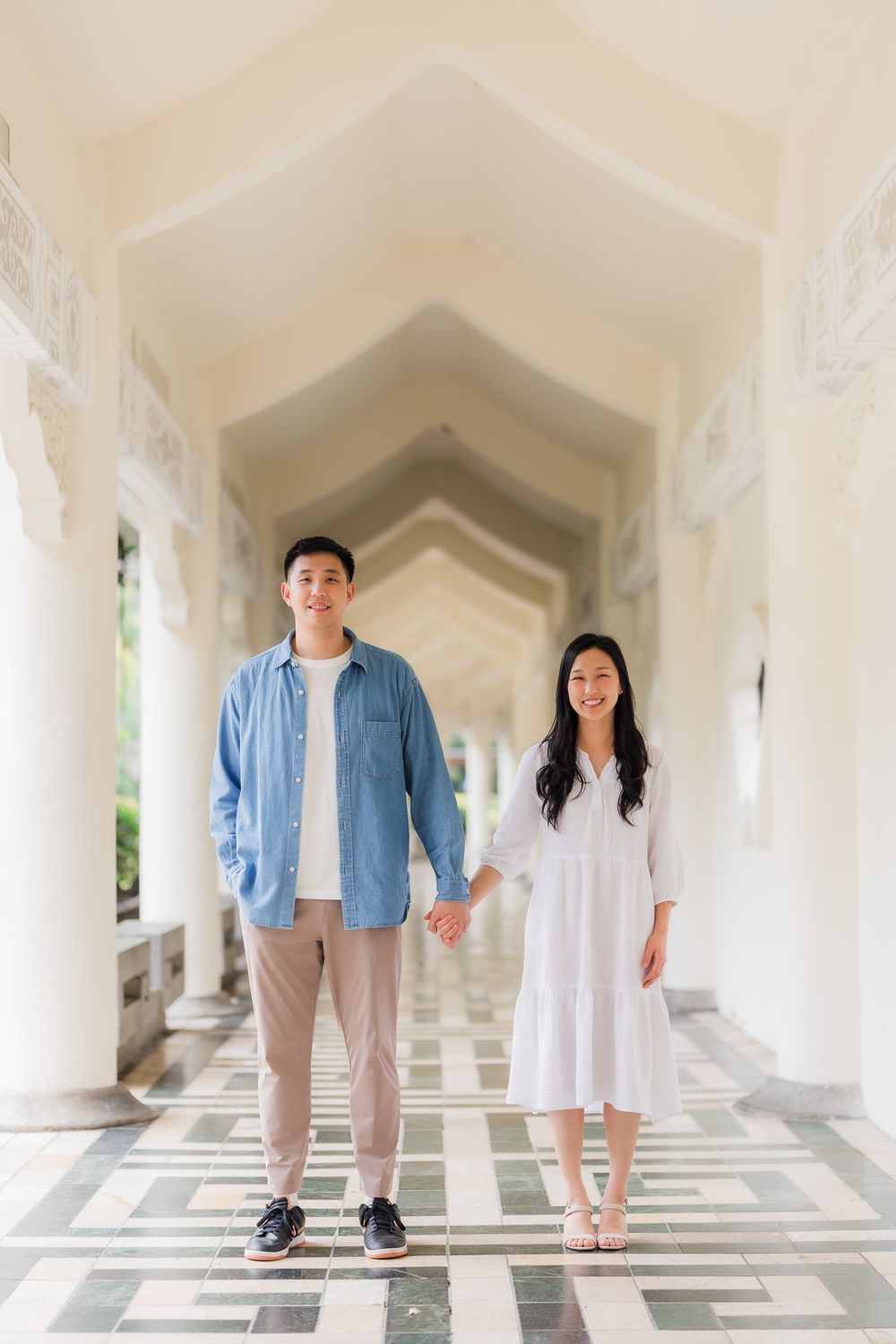 A couple stands holding hands in a white arched hallway with geometric floor patterns creating a romantic architectural setting.