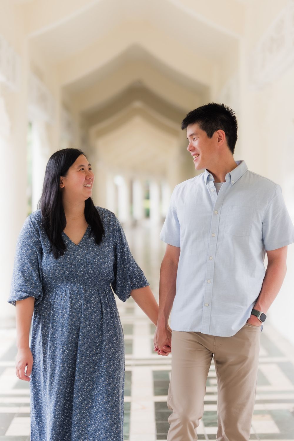 A couple in coordinated blue attire shares a joyful smile at each other while walking through a bright, white-columned corridor.