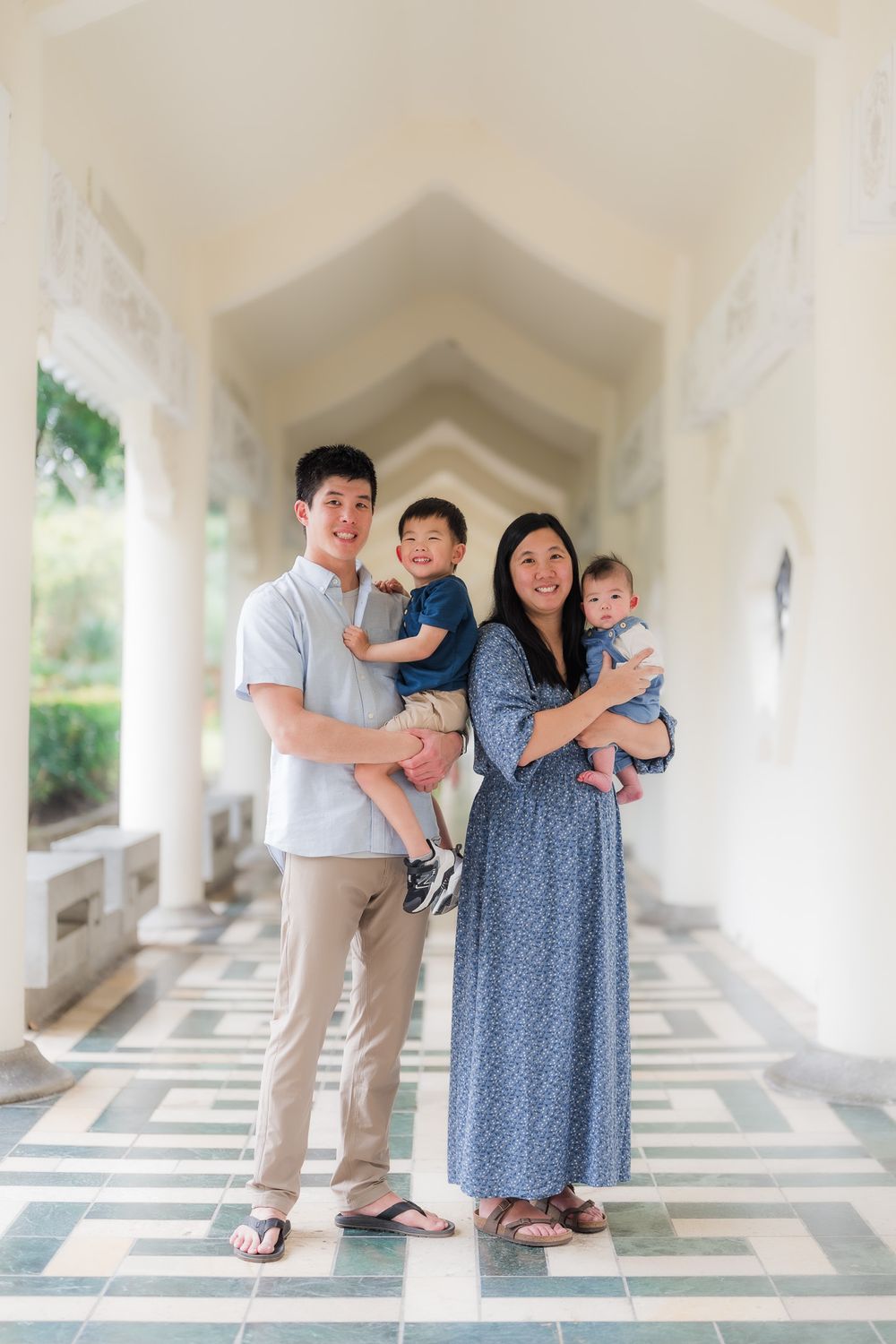 Family in coordinated blue and neutral outfits poses together in an elegant white archway corridor with geometric style floor tiles.