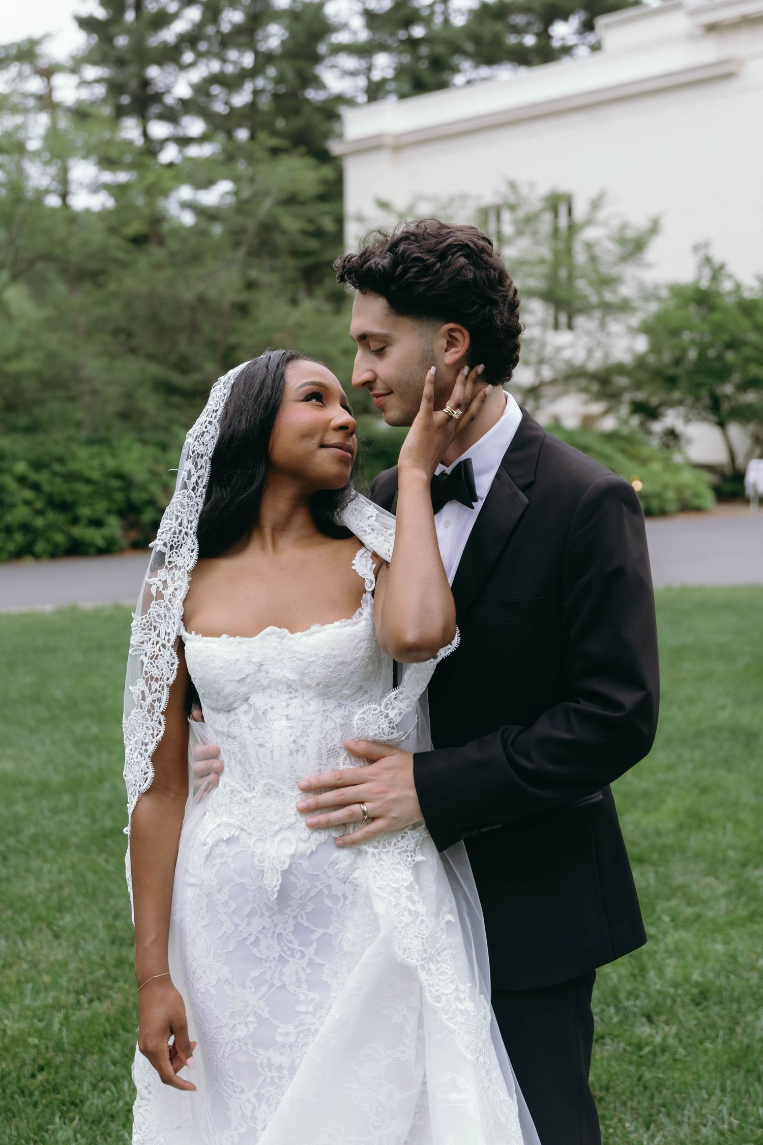 Romantic wedding couple embraces outdoors on grassy lawn with white building and evergreen trees in background.