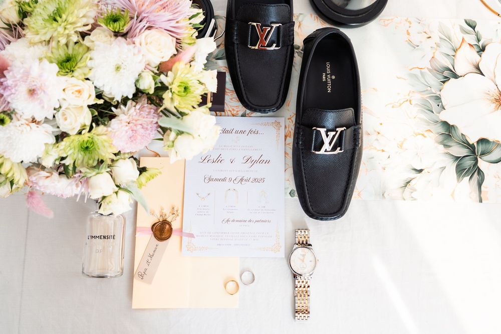 Wedding details flatlay with Louis Vuitton black loafers, invitation, rings, watch and pink floral arrangements.