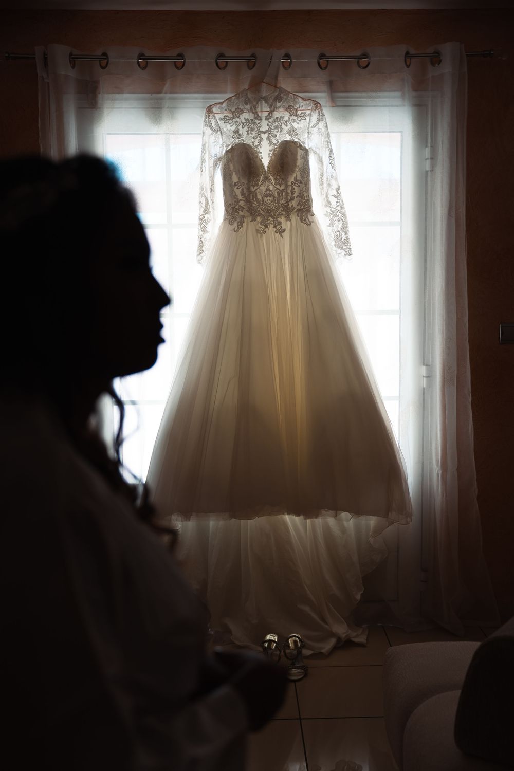 A white lace wedding gown hangs elegantly in front of a sunlit window.