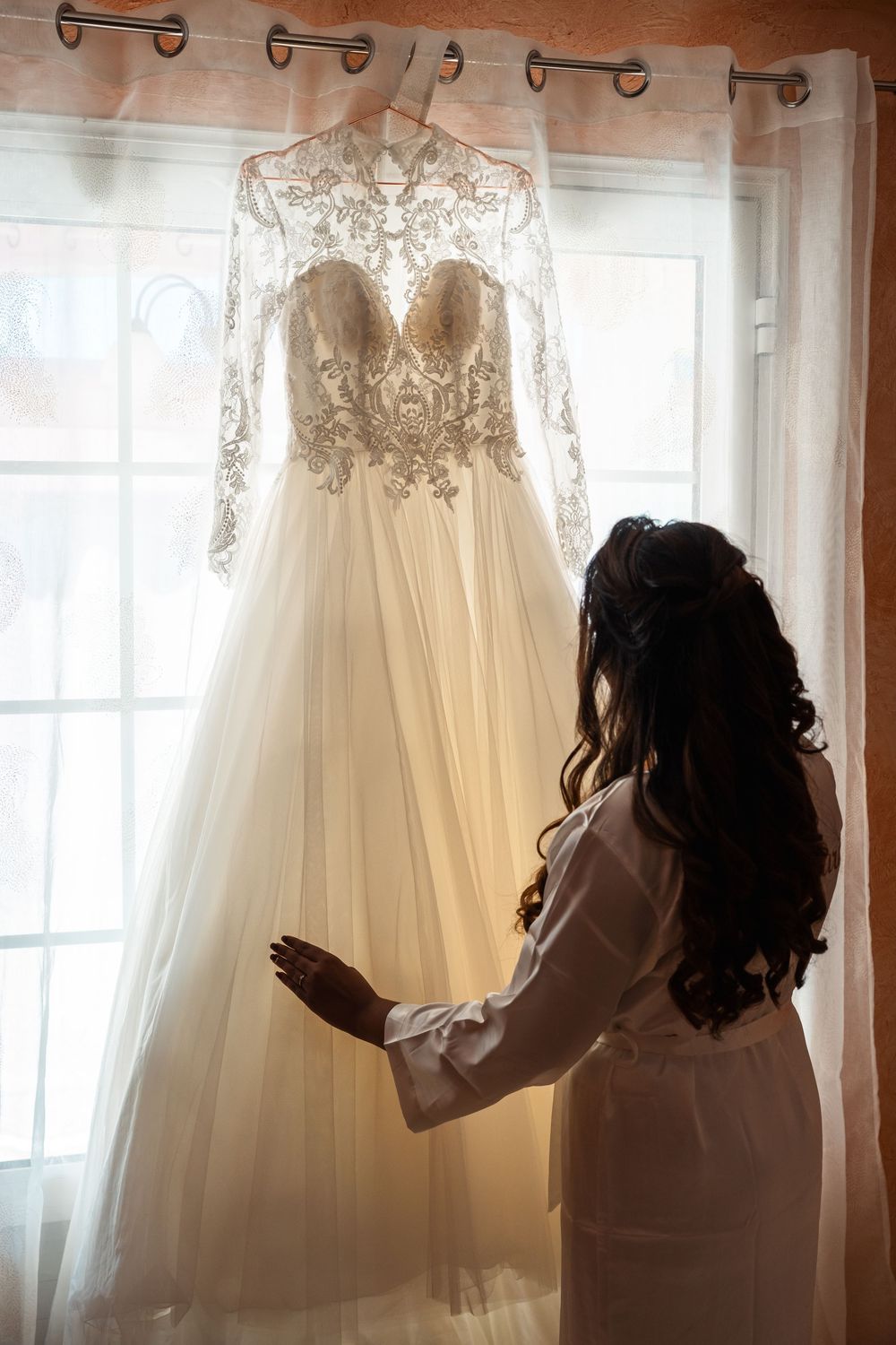 Silhouettes of people admiring a wedding dress hanging by a window in soft natural light.