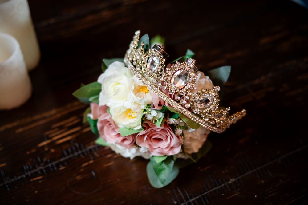 A delicate gold crown adorned with jewels sits elegantly beside pink and white flowers on a dark wooden surface.
