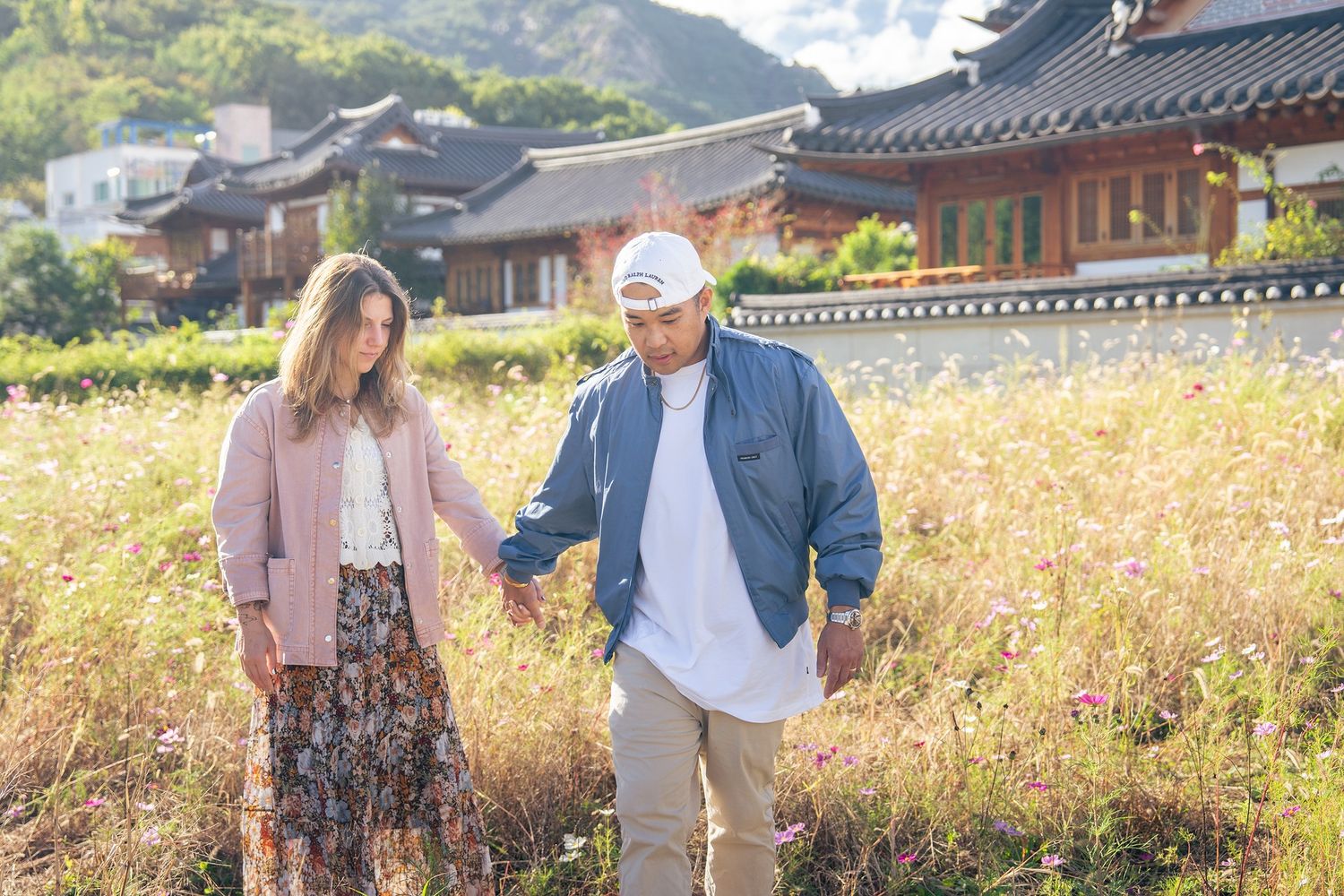 People stroll through a scenic autumn garden path with traditional Korean architecture and mountains in the background.