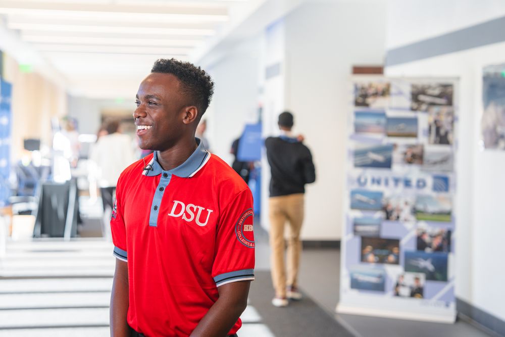A student in a red DSTU polo shirt smiles in a bright university hallway.