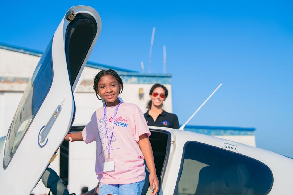 Young girl smiles near a small aircraft with a female pilot instructor behind her on a sunny day.