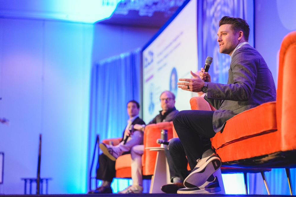 Three professionals engaged in a panel discussion on stage with orange chairs and blue lighting backdrop.