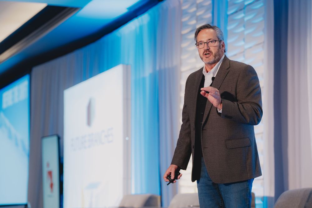 Professional male speaker presenting on stage at a corporate conference with blue backdrop and podium.