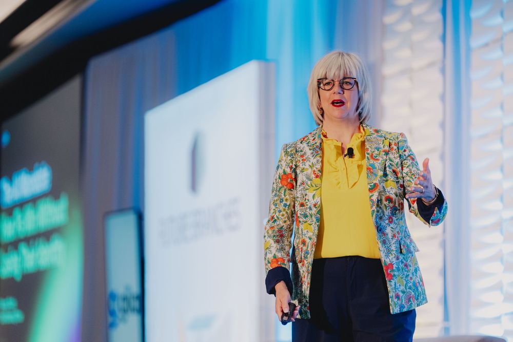 Woman speaker in floral blazer presenting on stage at a professional conference with blue backdrop.