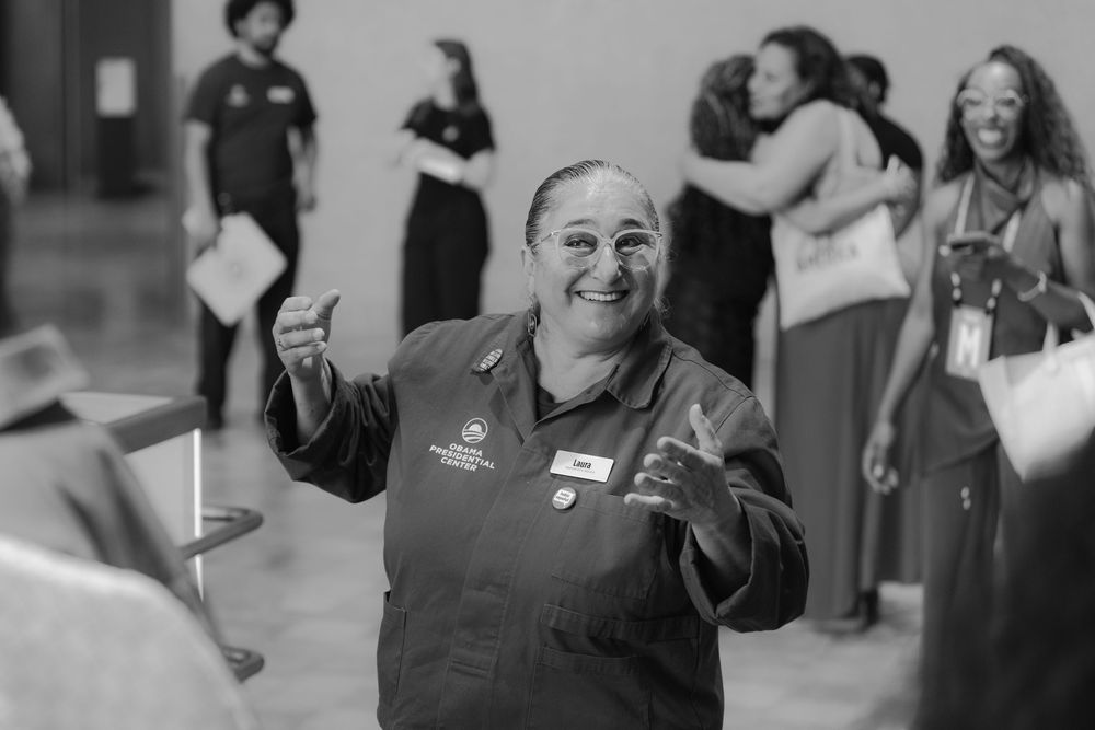 Smiling woman in uniform giving thumbs up at an indoor event with people socializing in the background.