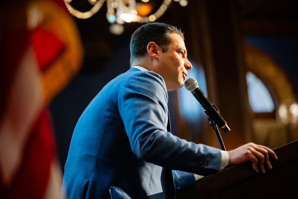 Man in blue suit speaking at podium with microphone at formal indoor event.