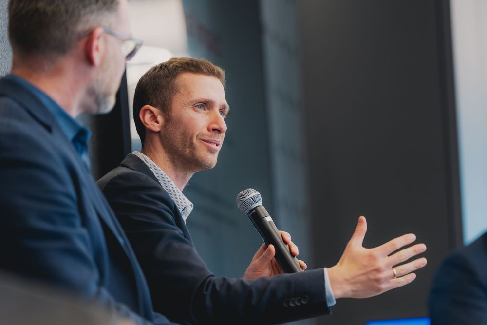 Two businessmen in suits at a panel discussion, one speaking into a microphone while gesturing with his hand.