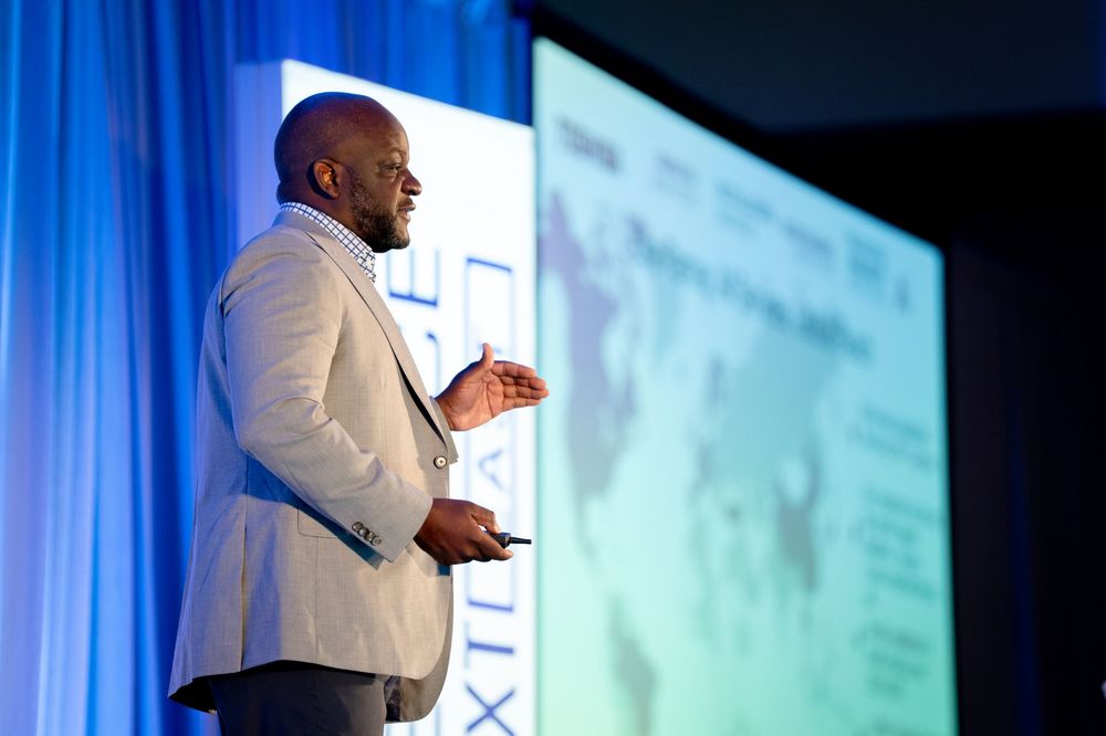 Black male speaker in white blazer presents on stage at professional conference with blue backdrop.