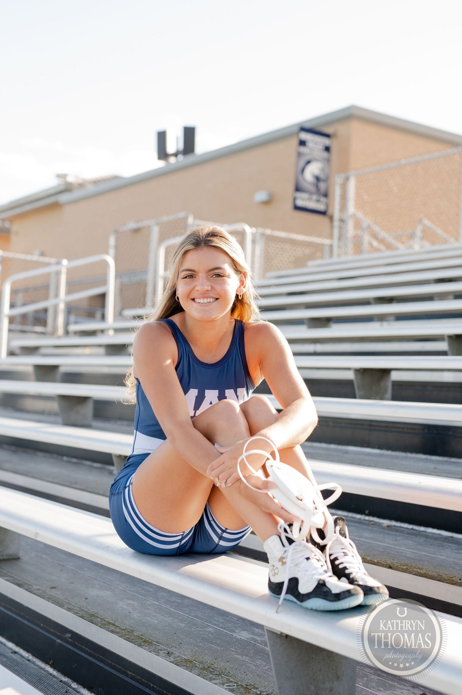 Athlete in blue uniform sits on bleachers tying white sneakers at a sports stadium.