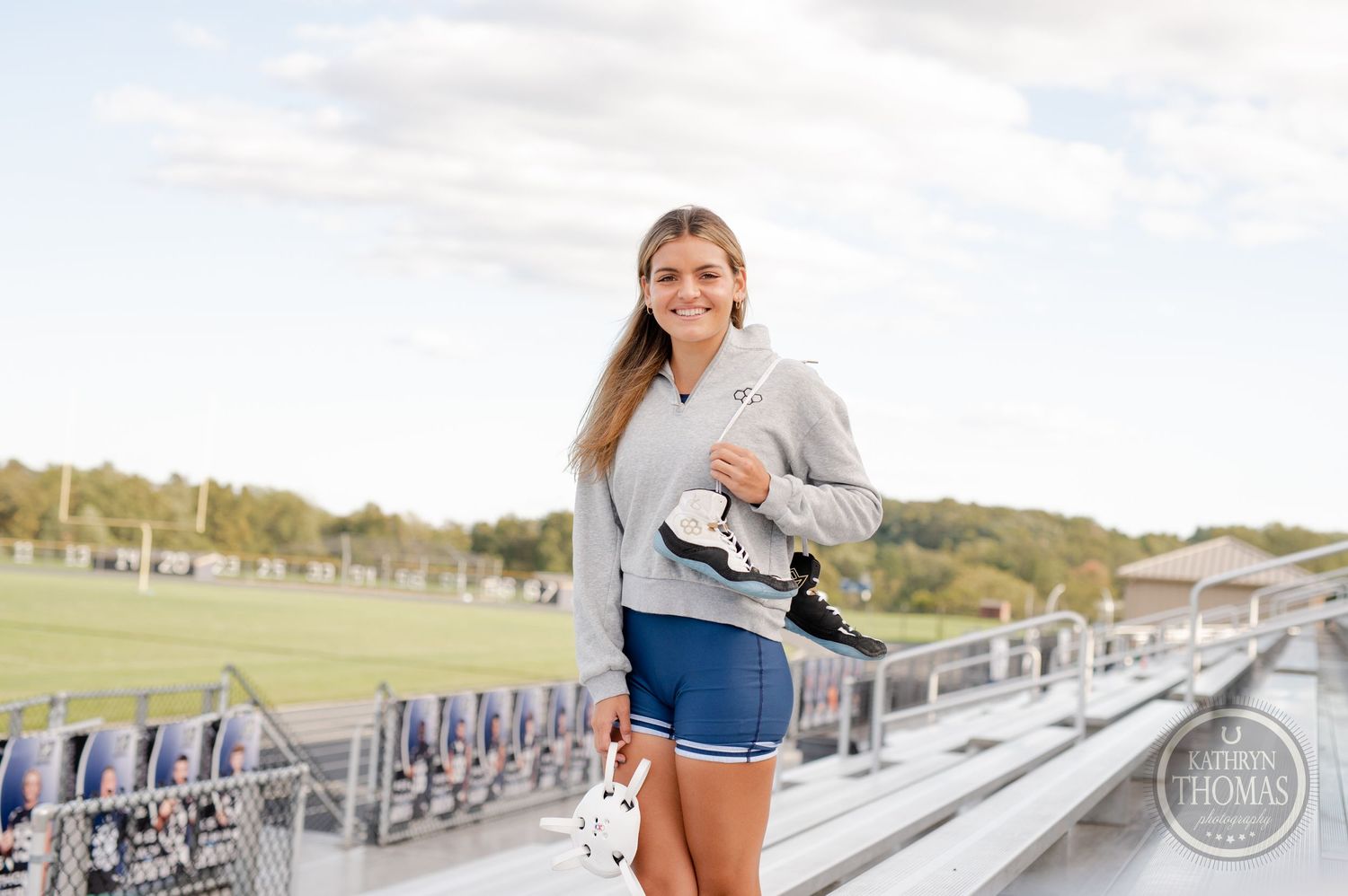 Athlete in grey sweatshirt and blue shorts standing on stadium bleachers holding sports equipment.