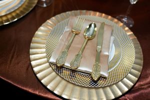 Elegant place setting with gold charger plate and silverware on brown tablecloth.