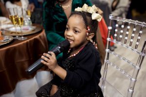 Young child in a black dress holding a microphone and smiling during a celebration.