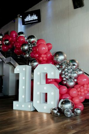 Illuminated number 16 display with red and silver balloon arch decoration against white wall.
