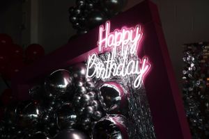 Neon pink 'Happy Birthday' sign glowing against dark balloons in party decoration.