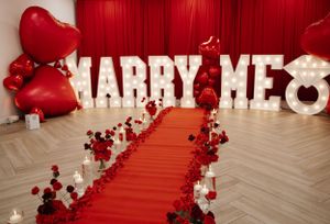 Red carpet proposal setup with 'MARRY ME' in light-up letters and heart-shaped balloons.