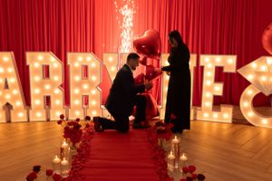 Romantic proposal scene with illuminated 'MARRY ME' letters and red carpet lined with candles.