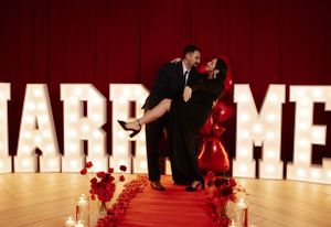 Romantic moment captured in front of illuminated 'MARRY ME' sign on red carpet.