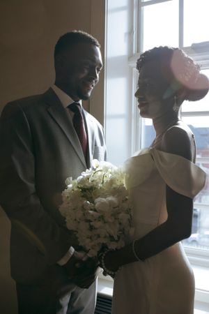 Bride and groom share an intimate moment by a window with white bouquet.